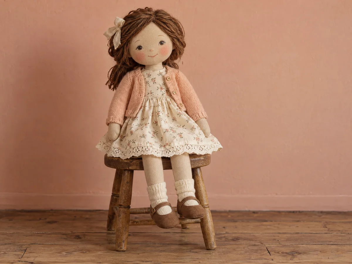 A brown-haired cloth doll in a cream dress and peach cardigan seated on a wooden stool